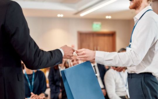 A young man in a button-down white shirt and slacks hands a giveaway bag to a fellow tech conference attendee.