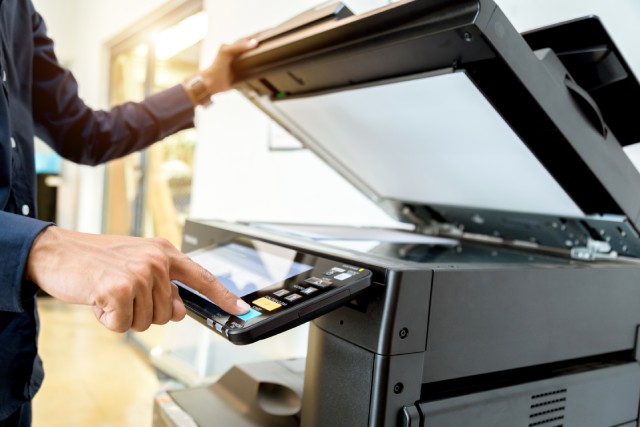 An office copy machine with a business member standing at the trackpad to create a copy order. The lid is open on the machine.