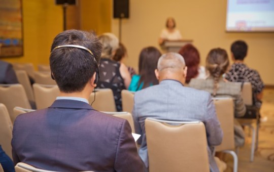 A person wearing headphones while sitting in a conference room as a speaker stands at a podium and gives a presentation.