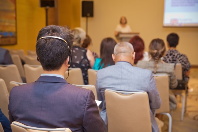 A person wearing headphones while sitting in a conference room as a speaker stands at a podium and gives a presentation.