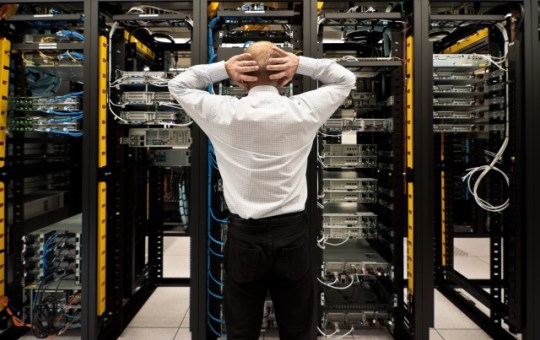 A man stands in front of server racks that hold servers and cables. His hands are behind his head as he gazes at the setup.