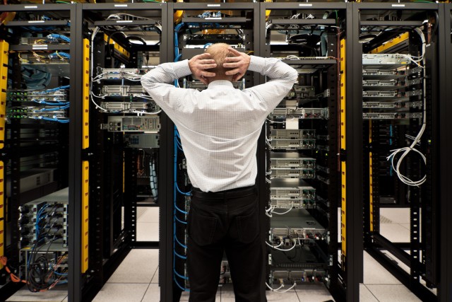 A man stands in front of server racks that hold servers and cables. His hands are behind his head as he gazes at the setup.