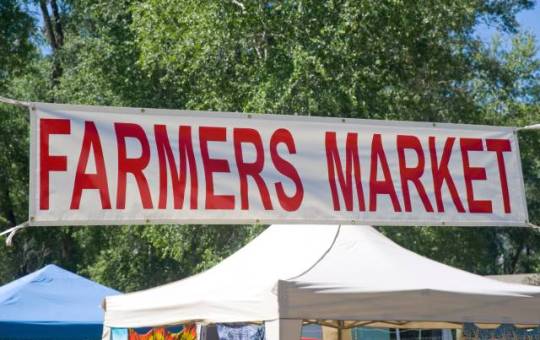 A very large, white banner that reads "Farmers Market" in big, red letters stretched in front of trees and tents.