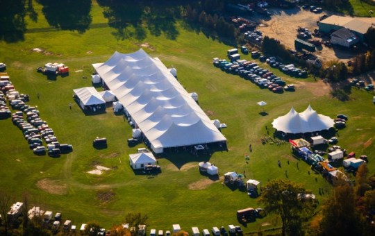Aerial view of an outdoor event with several large event tents set up in the grass and a parking area for cars.