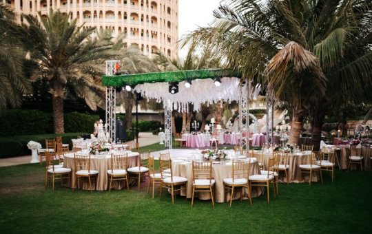 Event tables sit near a temporary dance floor and stage for an outdoor wedding event in a park near palm trees.