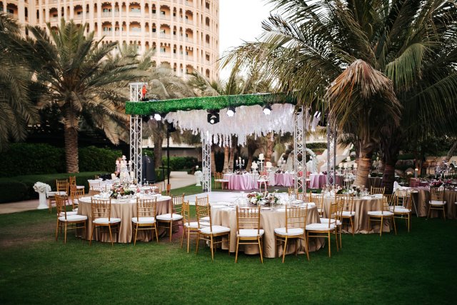 Event tables sit near a temporary dance floor and stage for an outdoor wedding event in a park near palm trees.