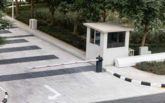 A security booth with a car barrier guards the entryway into the premises. The booth is compact and near some trees.