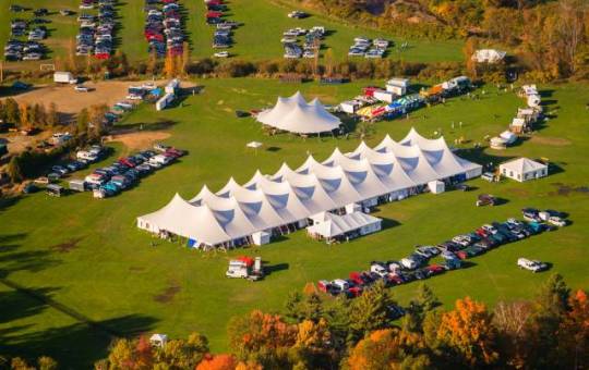 An aerial view of a large outdoor event featuring a central white tent, surrounded by smaller tents and guest cars.