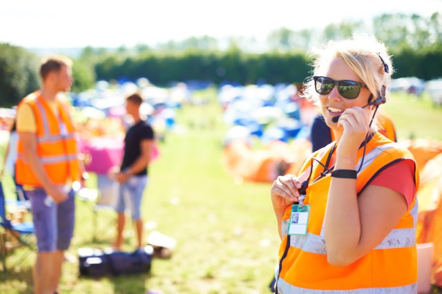 A blonde-haired woman wearing an orange high-visibility vest stands outside while talking into a headset.