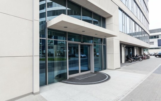 The exterior of a gray commercial office building features automatic sliding glass doors. The glass is tinted blue.