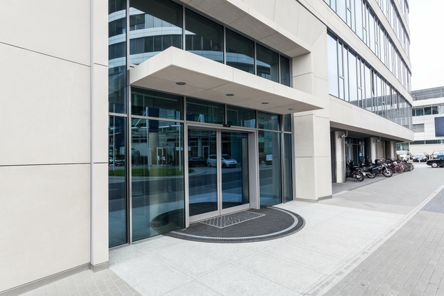 The exterior of a gray commercial office building features automatic sliding glass doors. The glass is tinted blue.