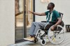 A man wearing pants and a green shirt in a wheelchair uses one hand to open a glass door to go inside a building.