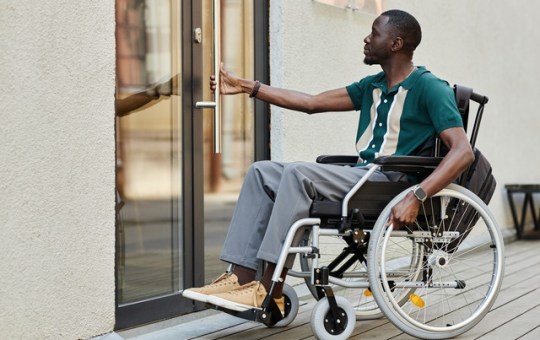 A man wearing pants and a green shirt in a wheelchair uses one hand to open a glass door to go inside a building.