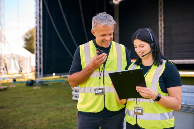Two production staff coordinate an outdoor event using a two-way radio, headsets, and a production plan.