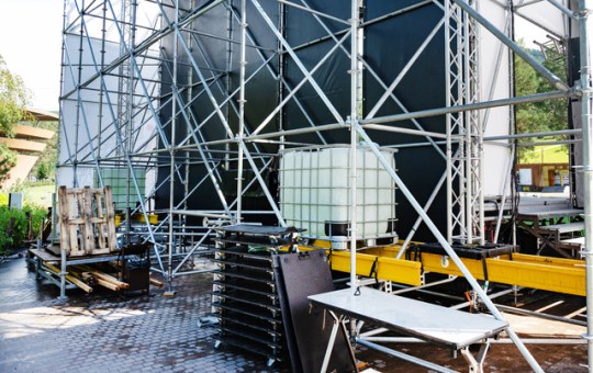 A metal scaffolding structure that’s been set up outdoors for an event with tables and pallets sitting around it.