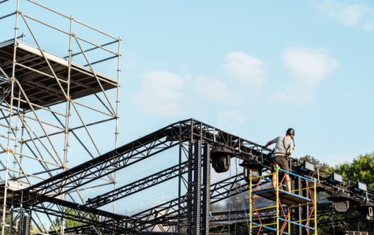 A person building a stage fixture for an outdoor event. He is standing on a tall, stable scaffolding structure.