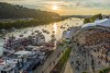 An aerial view of a large crowd enjoying a summer event near the banks of a calm river at sunset. Boats float on the water.